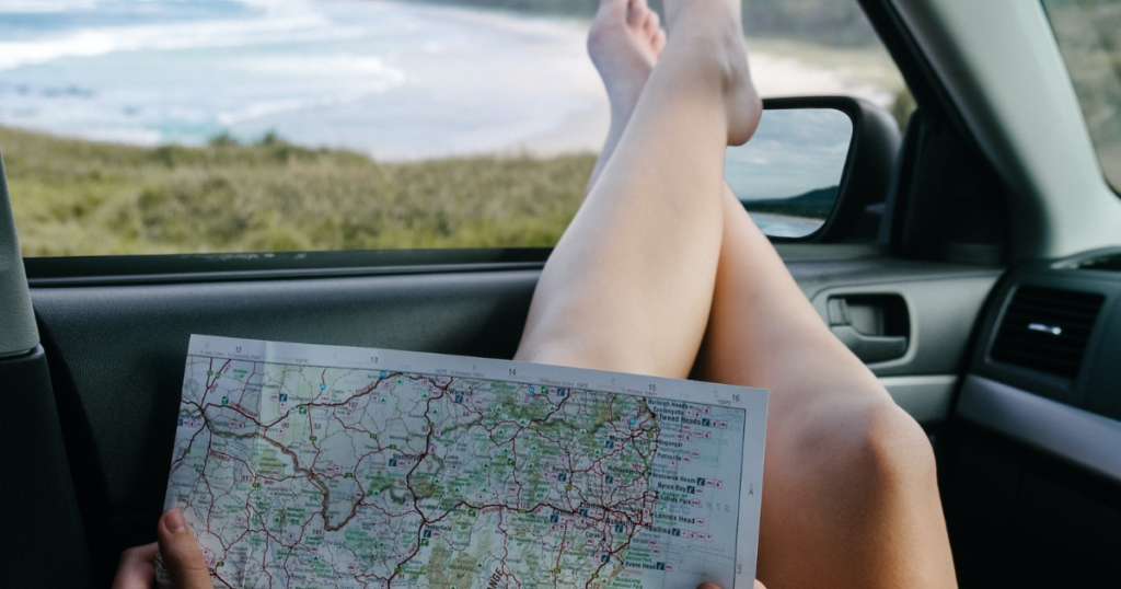 sitting inside a car, legs out of the window and a map on the lap. outside the car window a beach and water with waves 
