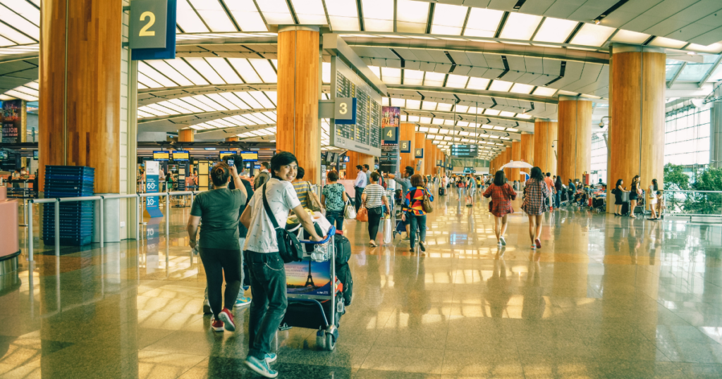 People in the airport. Guy is starting his world trip and carrying a luggage trolley through the hall and smiles to the camera. 