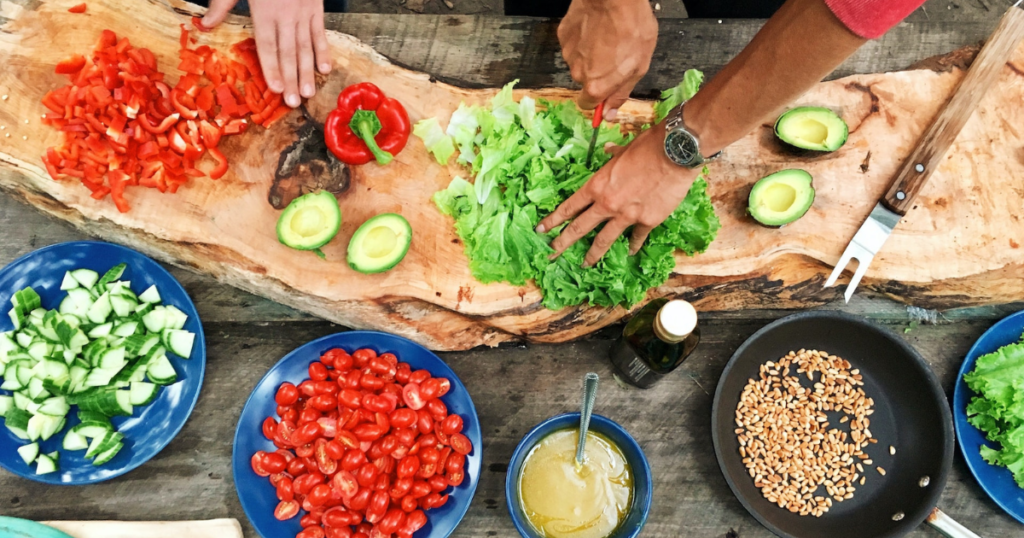 Cooking: a wooden table with lots of food like tomatos an salat, avocado and cucumbre. Hand on the picture cutting the vegetables