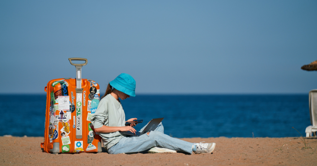 girl with big suitase ist sitting on the beach with laptop and smartphone, completely dressed. This picture should symbolize how helpful the best travel hacks can be. 