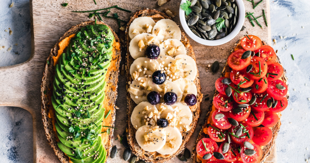 Slices of bread with vegan toppings like hummus, avocado, banana, tomato, pumpkin seeds, blueberrys. Looks like a healthy and delicious vegan meal.